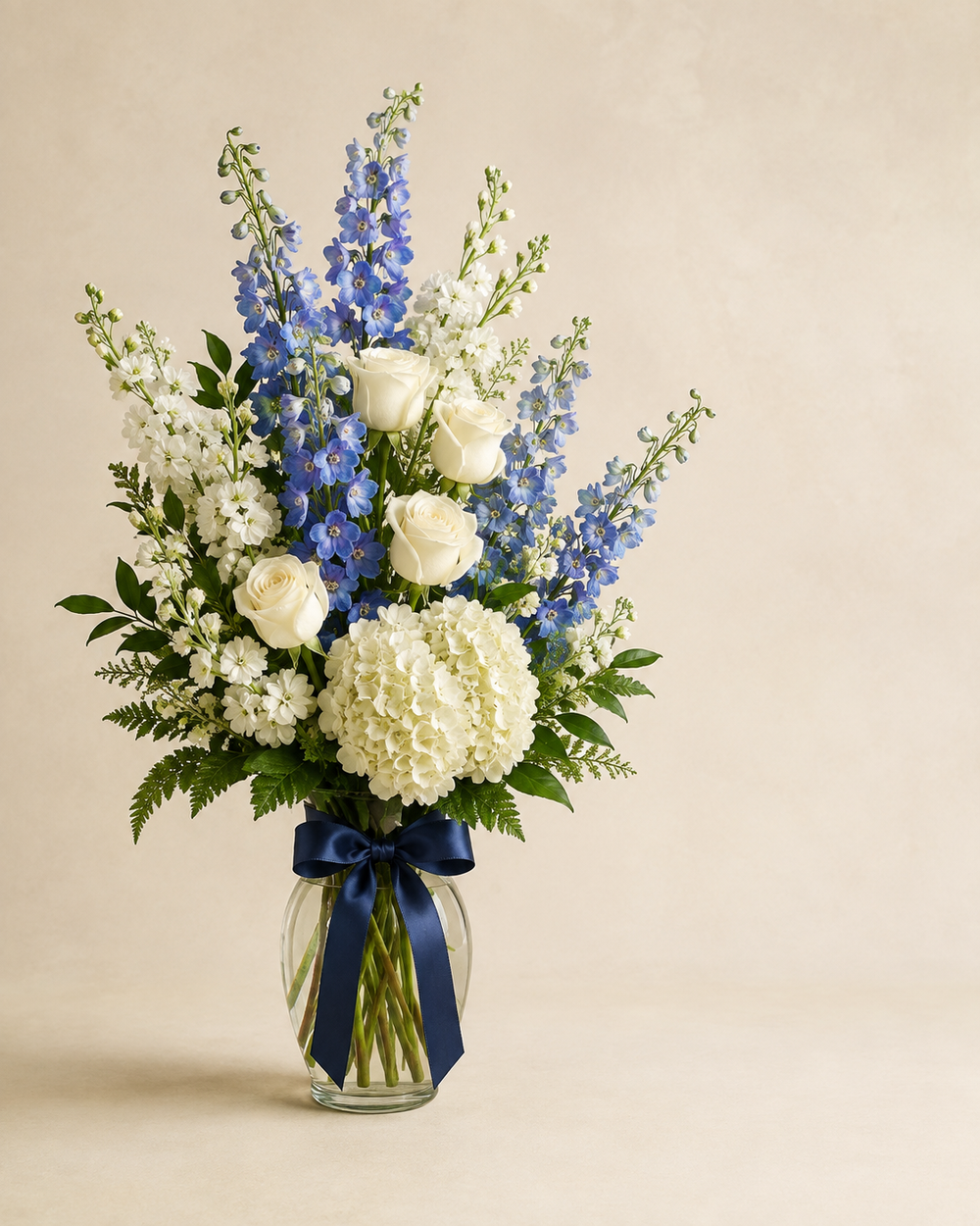Blue Quietude sympathy vase with white roses, blue delphinium, white hydrangeia, and white stock in a clear vase with navy ribbon