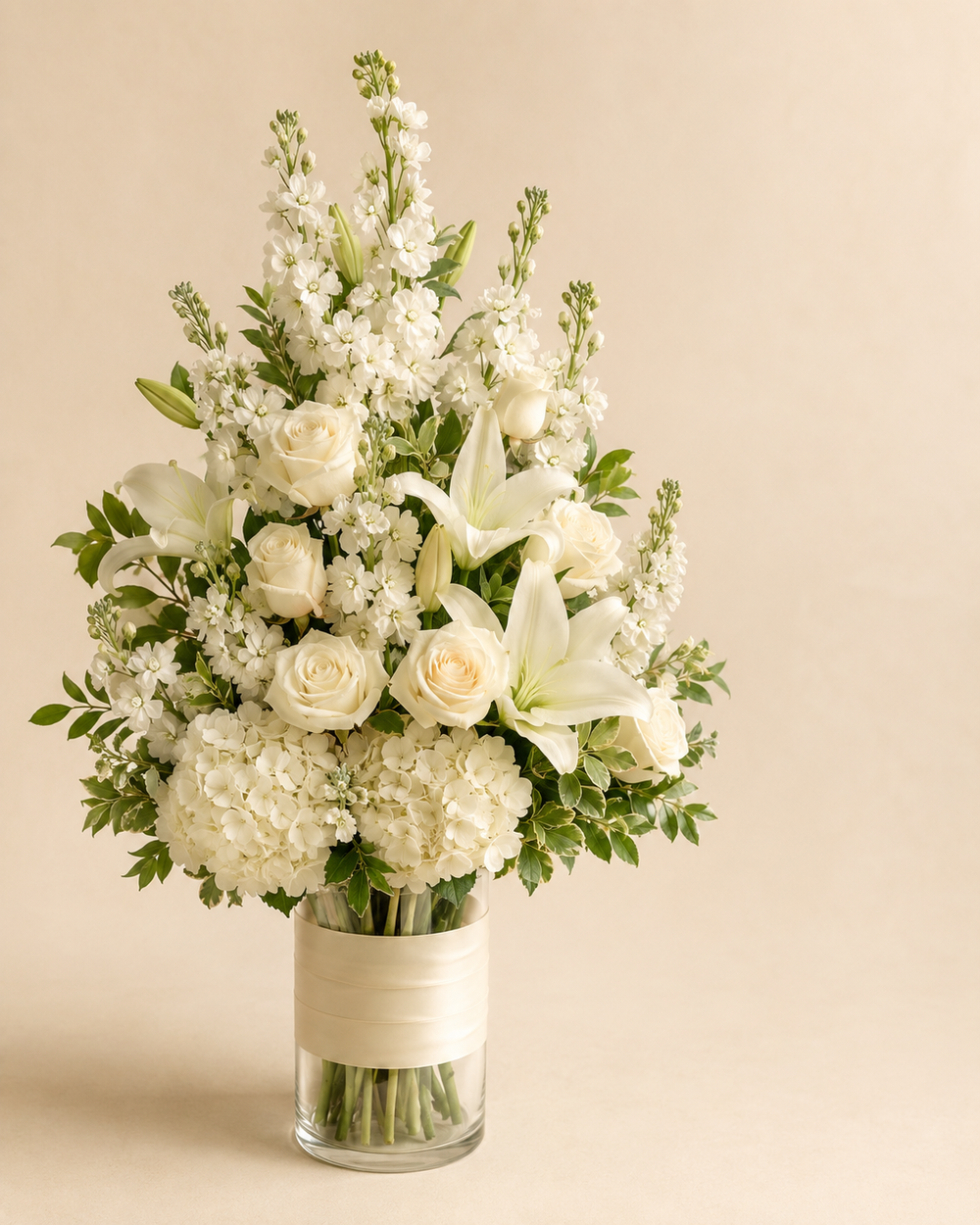 Ivory Lantern sympathy bouquet with white roses, Oriental lilies, hydrangeias, and stock in a clear cylinder with ivory wrap