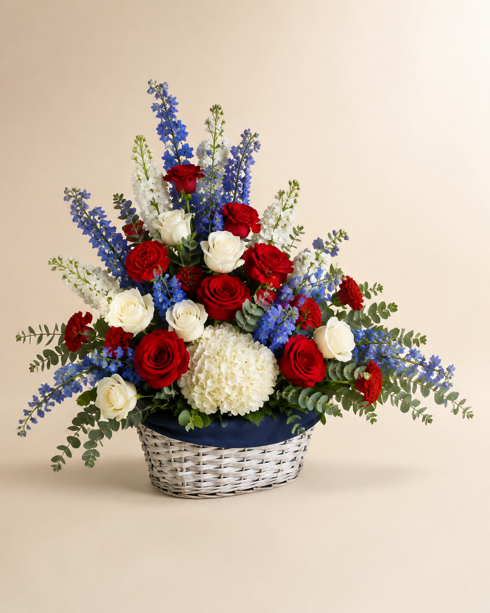 Liberty Garden sympathy basket with red and white roses, red carnations, white hydrangea and stock, blue delphinium, and eucalyptus in a navy-lined whitewashed basket