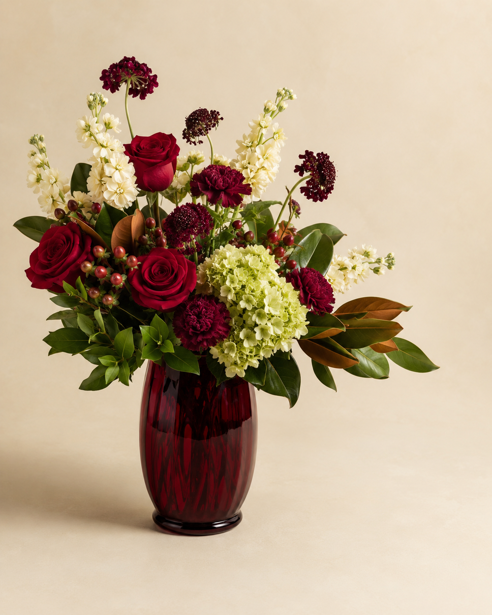 Ruby Orchard Memorial with deep-red roses, burgundy carnations and scabiosa, green hydrangea, cream stock, hypericum, and magnolia foliage in a dark ruby glass vase
