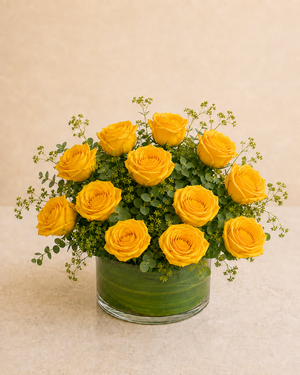 Exactly 12 yellow roses in a low leaf-wrapped clear glass vase